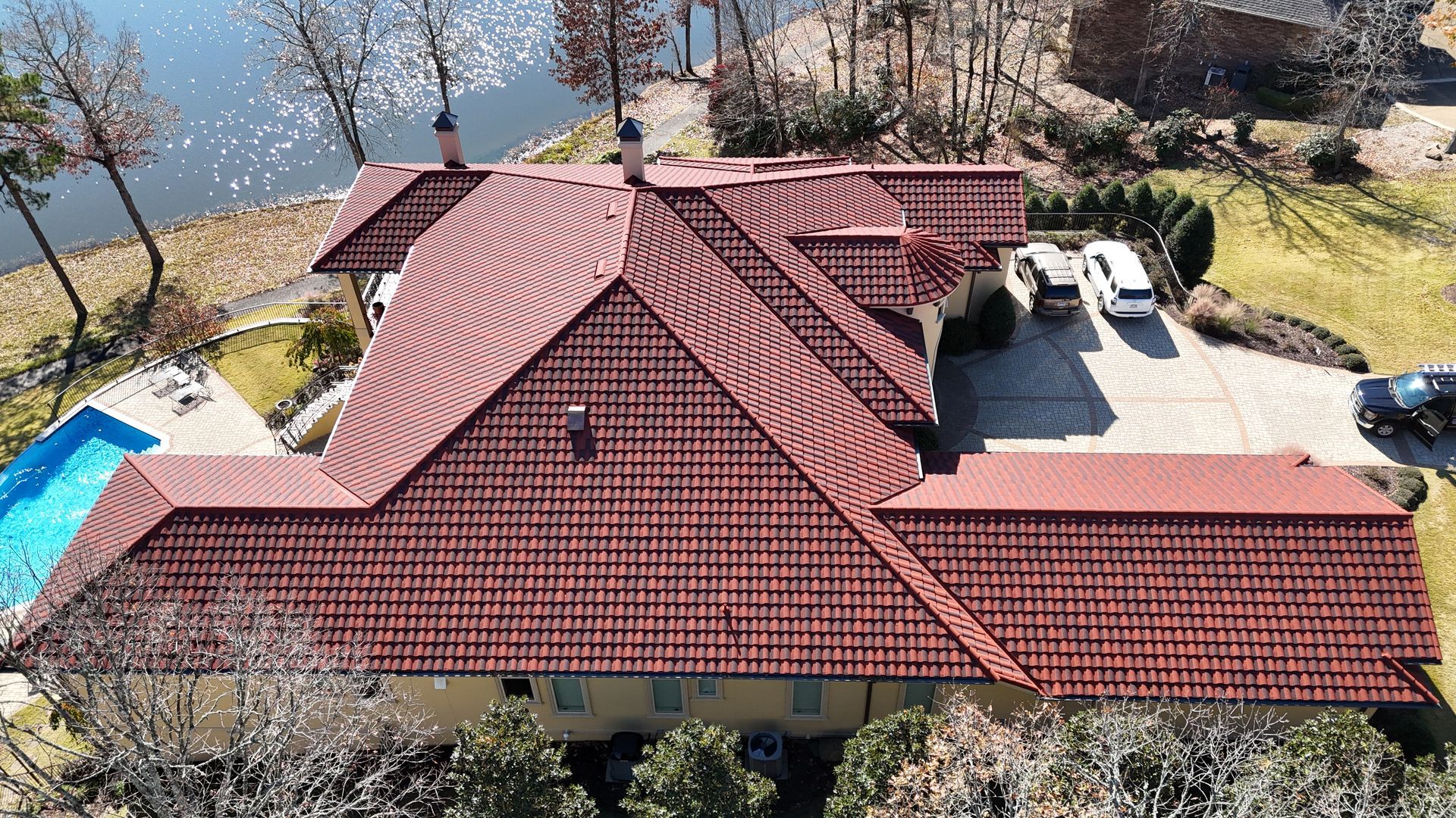 Overhead view of a house with a red-tiled roof, pool, cars in the driveway, and a lake in the background.