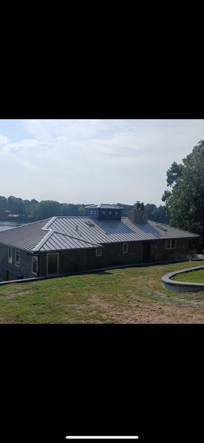 A long, single-story building with a dark metal roof. Green lawn in front, trees and sky in the background.