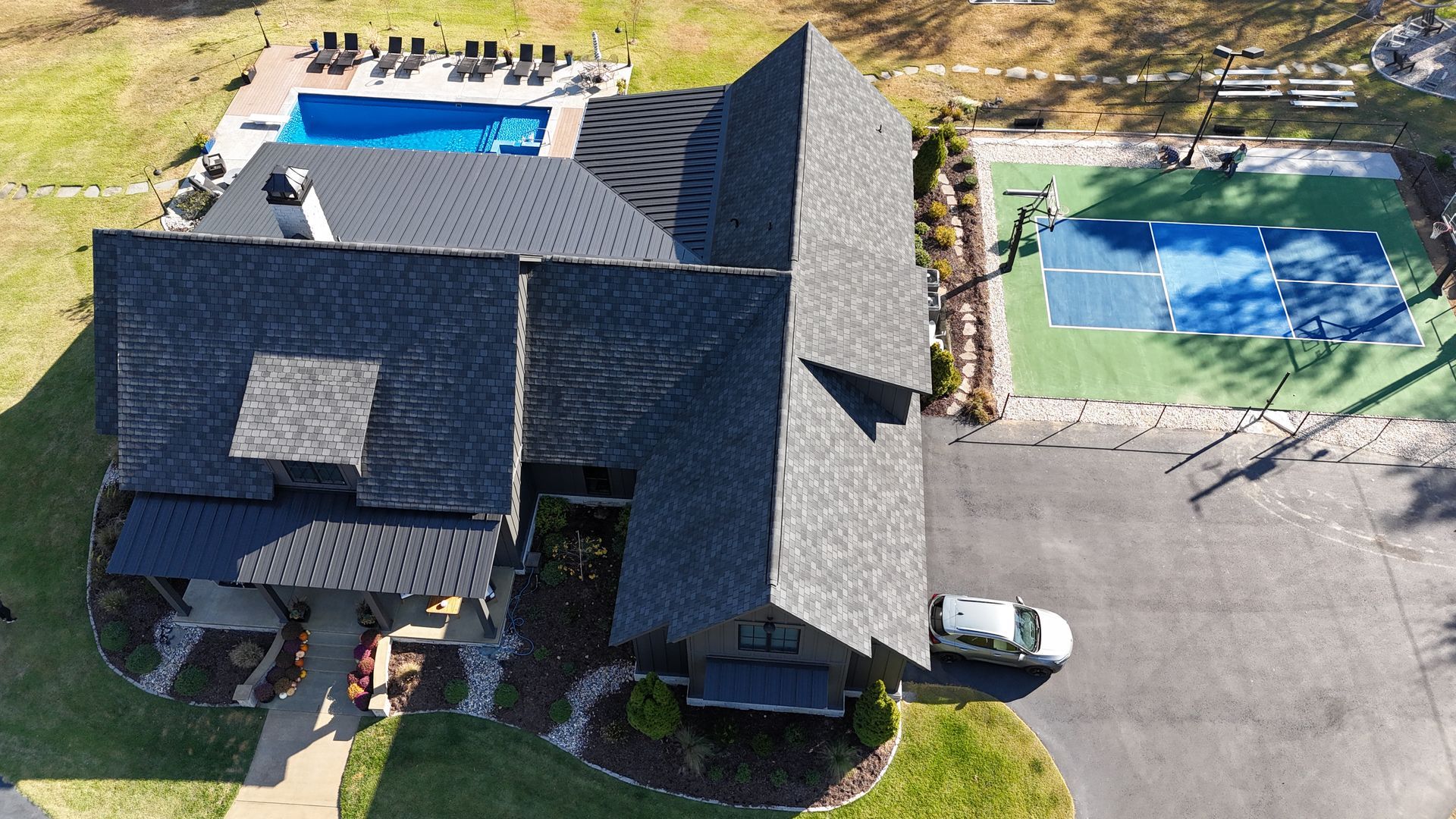 Overhead view of a house with a pool and tennis court. Dark roof, green lawn, and a white car parked in front.