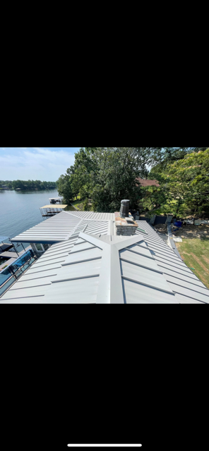 View of a metal roof of a lakeside home with a chimney, trees, and a lake in the background on a sunny day.