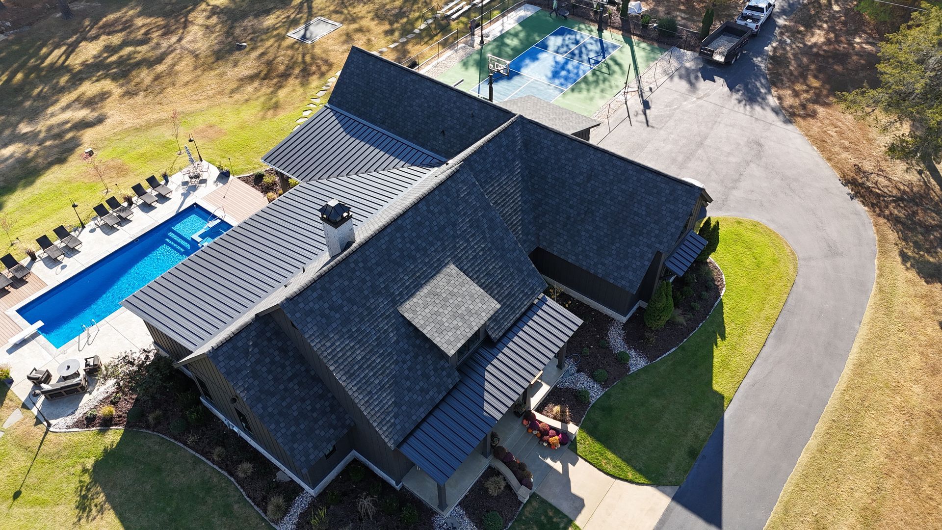 Aerial view of a dark-roofed house with a pool, driveway, and landscaping on a sunny day.