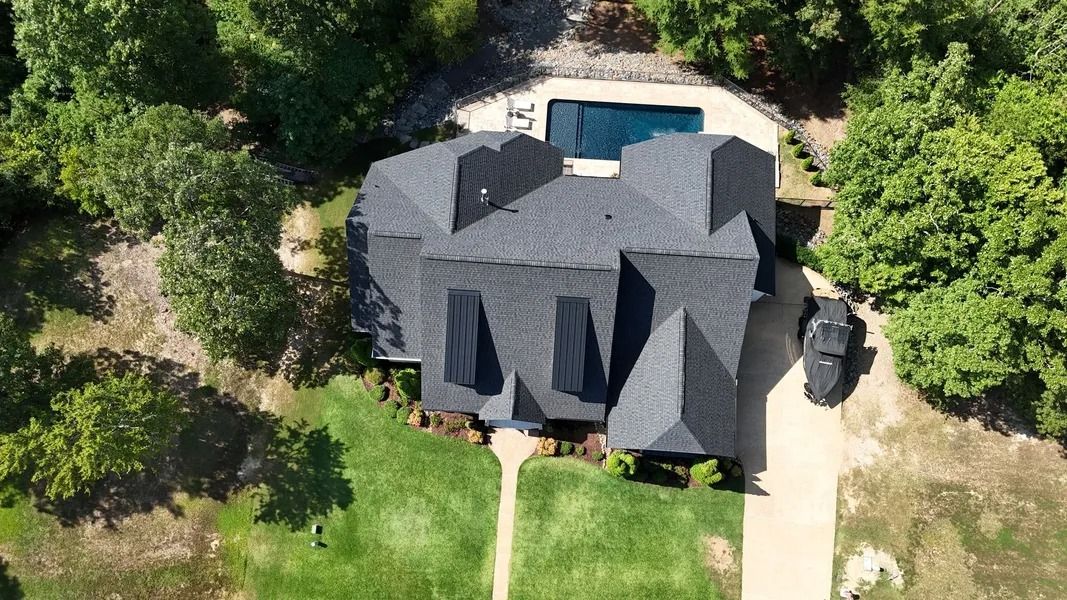 Aerial view of a gray-roofed house with a pool and driveway, surrounded by green trees and grass.