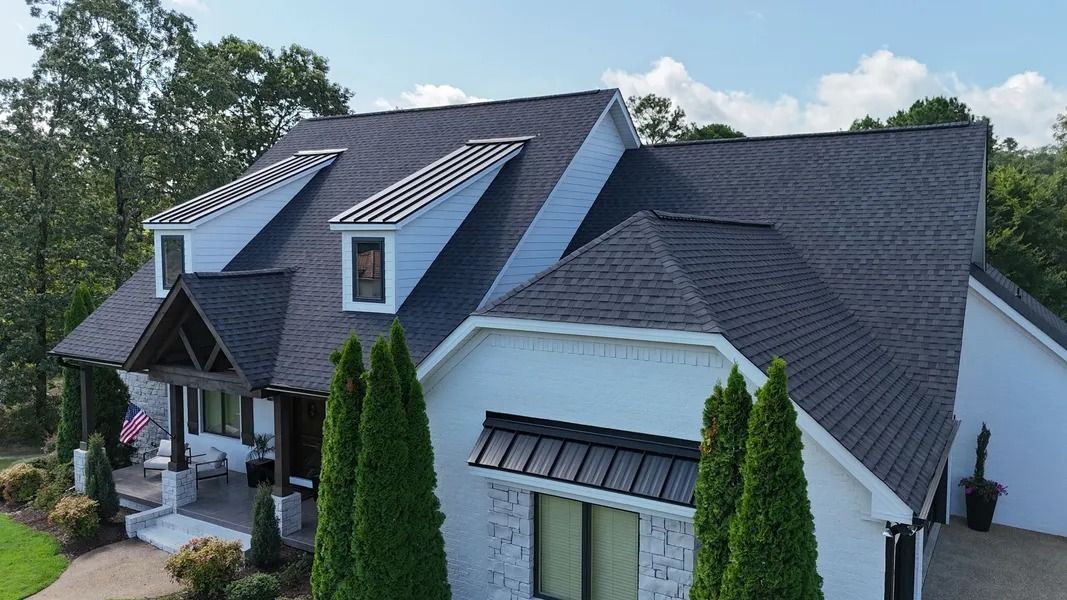 White house with a dark roof, featuring dormers and a porch, set against a blue sky.