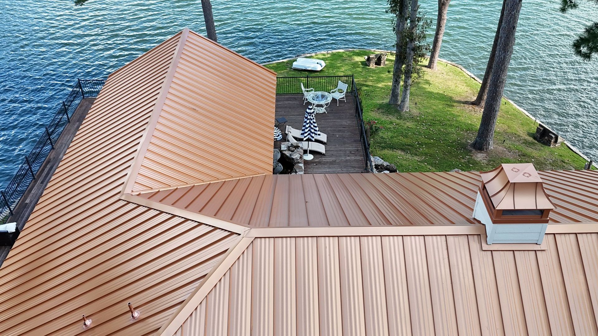 Copper-colored roof on a lake house, looking down from above. Lake in background. Steps lead to a grassy island.