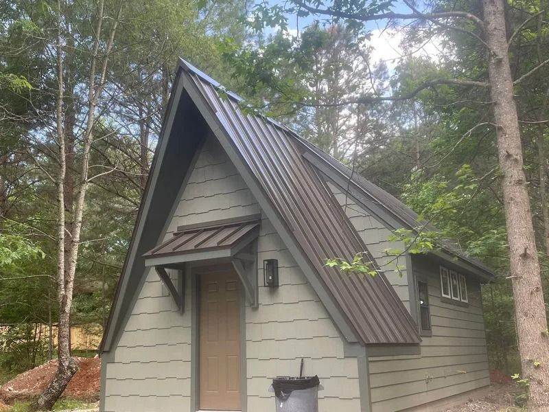 A-frame cabin with green siding, dark brown metal roof, and small awning in a wooded setting.