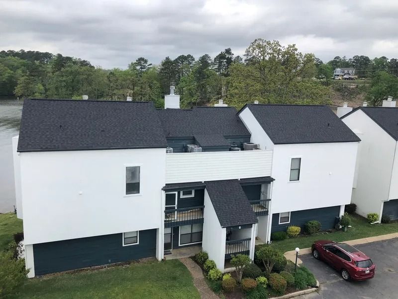 White townhouses with dark roofs near a lake and trees, a red car parked nearby.