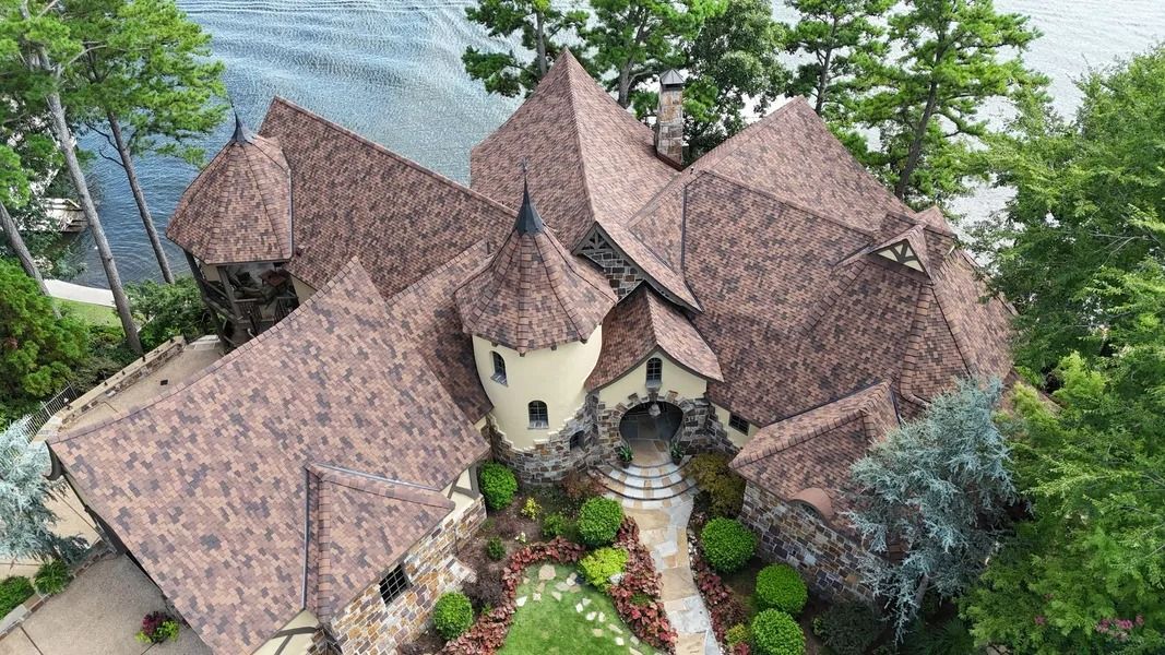 Overhead view of a large stone house with multiple gabled roofs, a tower, and a pathway leading to a lake.