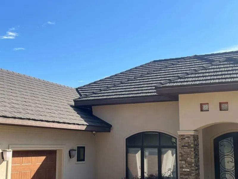A house with a brown tiled roof against a blue sky; beige walls, arched windows, and a wooden door.