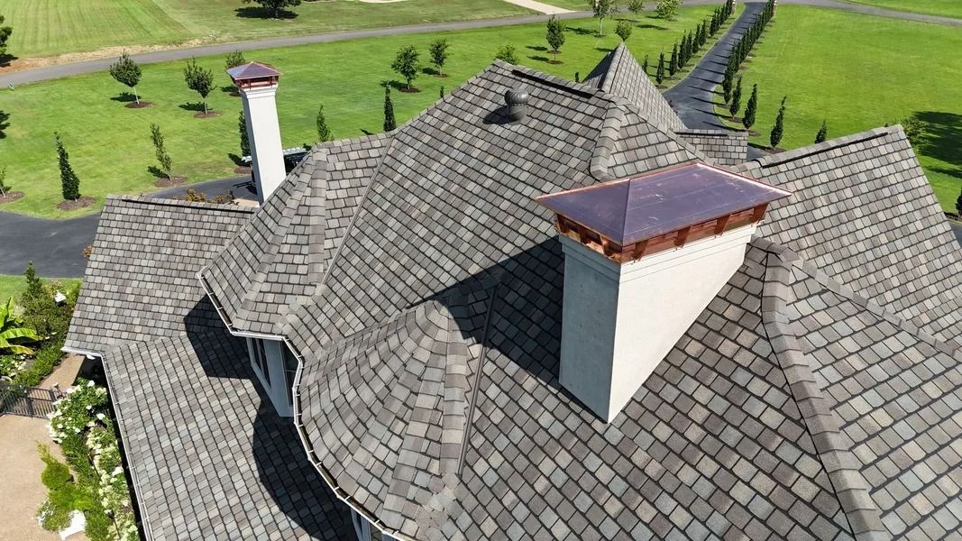 High-angle view of a home with a gray roof, chimneys, and a driveway lined with trees.