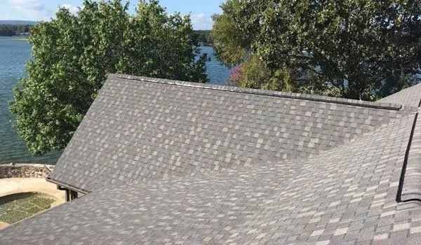 Asphalt shingle roof of a house with a water view and green trees in the background.