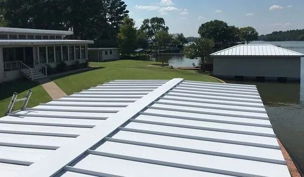 White dock on a lake with a house and boathouse in the background. Sunny day with trees and water.