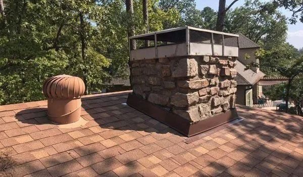 Chimney on a brown shingle roof with a cylindrical vent next to it, surrounded by trees.