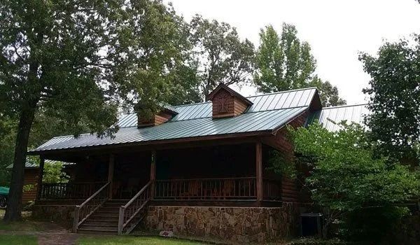 Wooden cabin with green metal roof, porch, steps, and two dormers amongst trees.