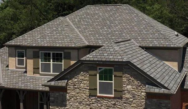 House with a gray shingle roof, stone facade, and green shutters against a sunny backdrop.