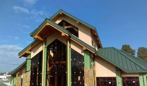 Building under construction with green roof, brown trim, beige walls, and dark glass windows against blue sky.
