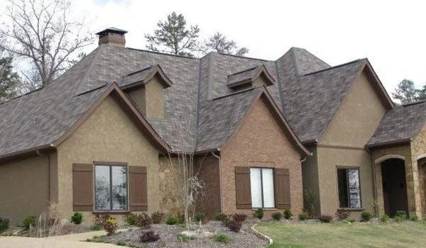 House with brown roof, tan stucco, brick accents, brown shutters, and landscaping.