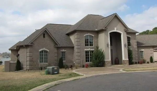 Large brick and stucco house on a sunny day with a circular driveway, bare yard, and blue sky.