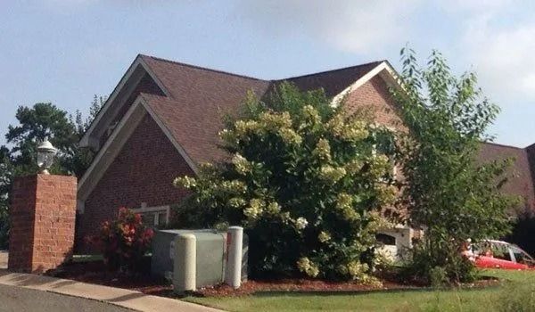 Brick house with brown roof and green bushes, electrical box in front yard.