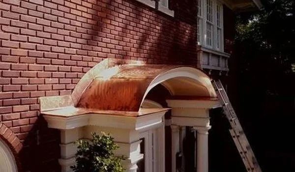 Copper-topped portico over a doorway on a brick building; a ladder leans against the building.