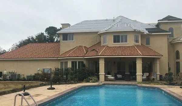 Large house with pool, tan stucco walls, red tile and gray shingle roofs, overcast sky.