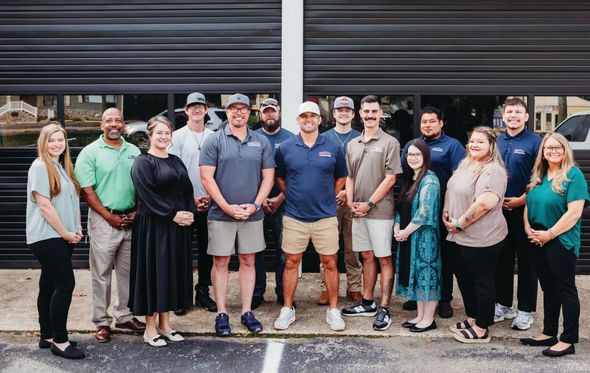 Group photo of 15 people in casual attire, standing in front of a gray garage door.