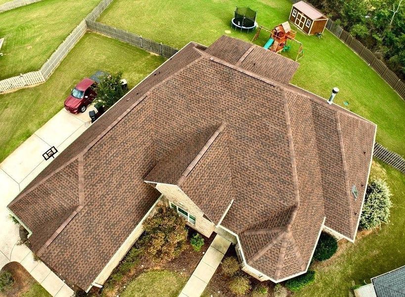 Aerial view of a large brown-roofed house with a driveway, lawn, and parked vehicles.