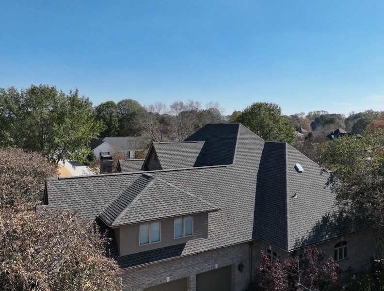 Suburban house with gray shingled roof beneath a clear blue sky, surrounded by trees.