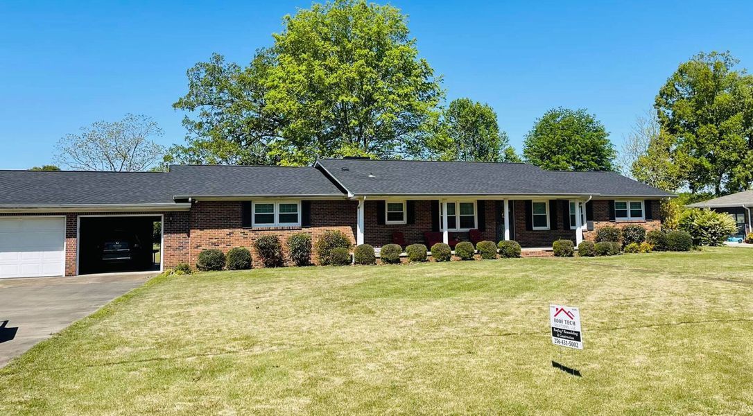 Single-story brick house with a large lawn, driveway, and trees under a clear blue sky