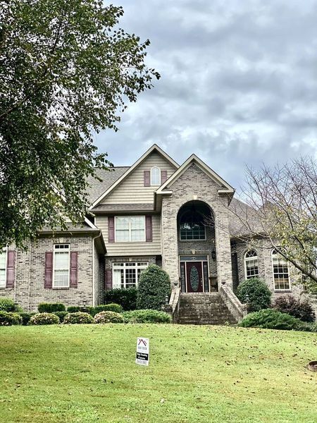 Two-story stone house with a front lawn, arched entryway, and trees framing the facade