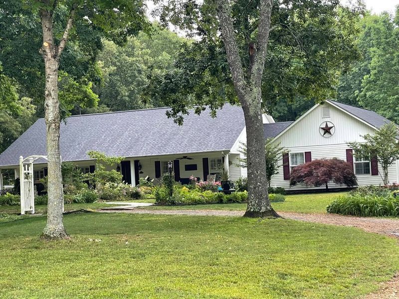 Single-story house with gray roof, white siding, front porch, and trees in a green yard