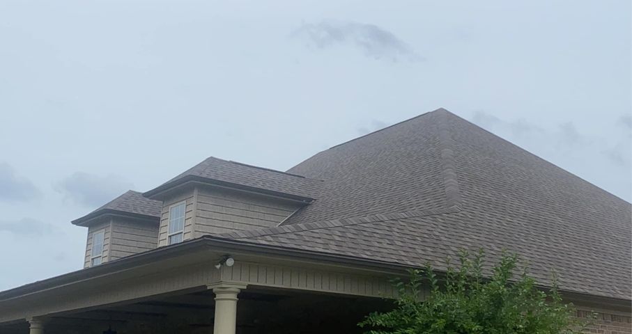 Large brown shingled house roof against a cloudy sky, with a small dormer and porch visible