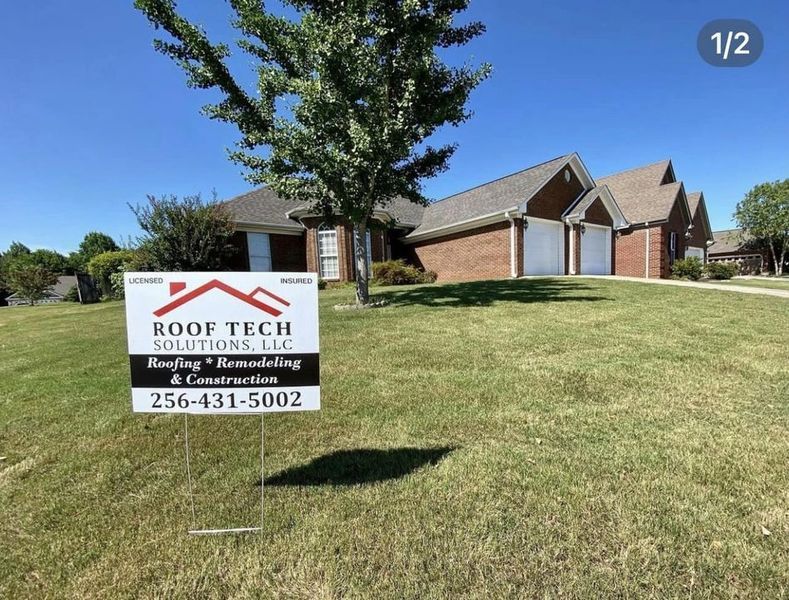 Roof Tech yard sign on a grassy lawn in front of a brick house under a clear blue sky