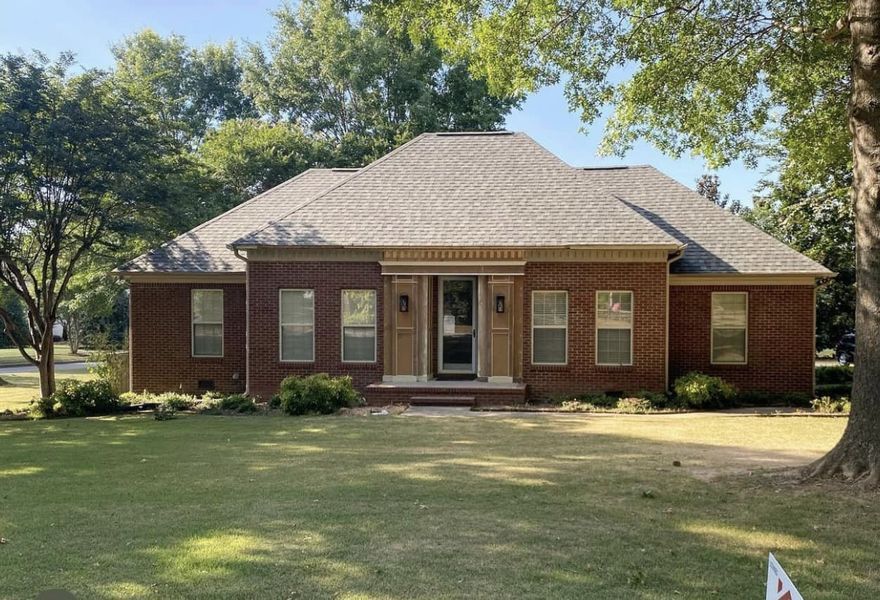 Single-story brick house with front porch and large lawn, framed by trees