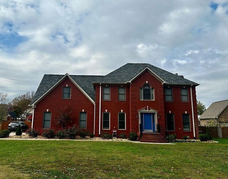 Red brick two-story house with a dark roof, blue front door, and large lawn under cloudy skies