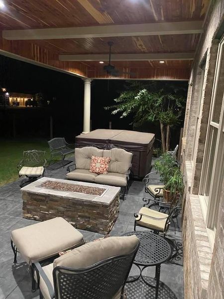 Covered patio with beige outdoor seating, stone fire pit table, and green yard at night