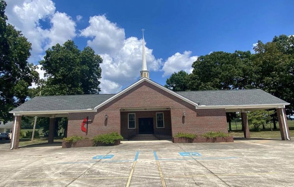Small brick building with covered side entrances, blue handicap parking spaces, and trees under a partly cloudy sky