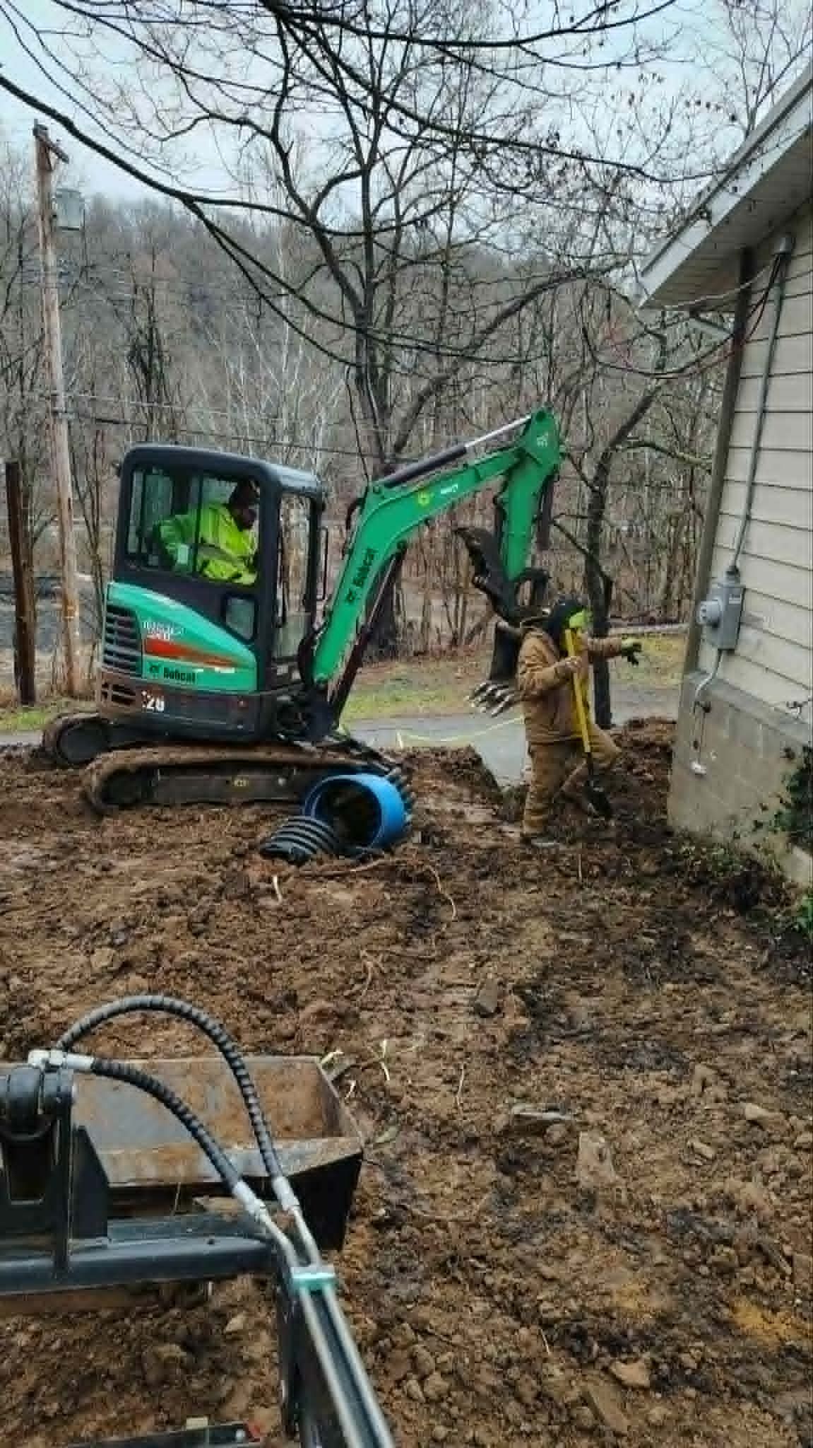 Mini excavator digging beside a house in a leaf-covered yard, with workers and tools nearby