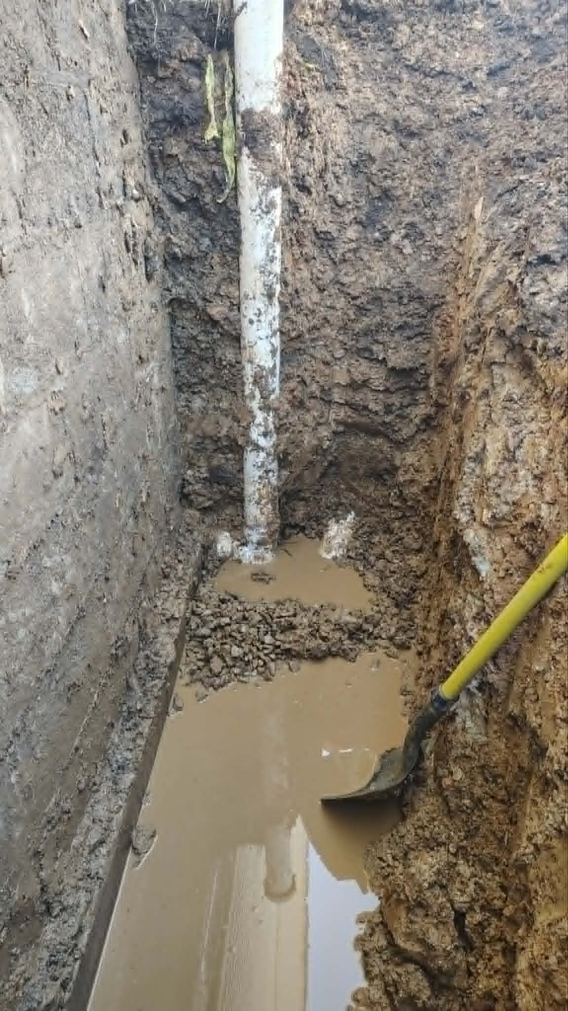 Narrow muddy trench with standing water and a yellow-handled shovel against exposed dirt walls