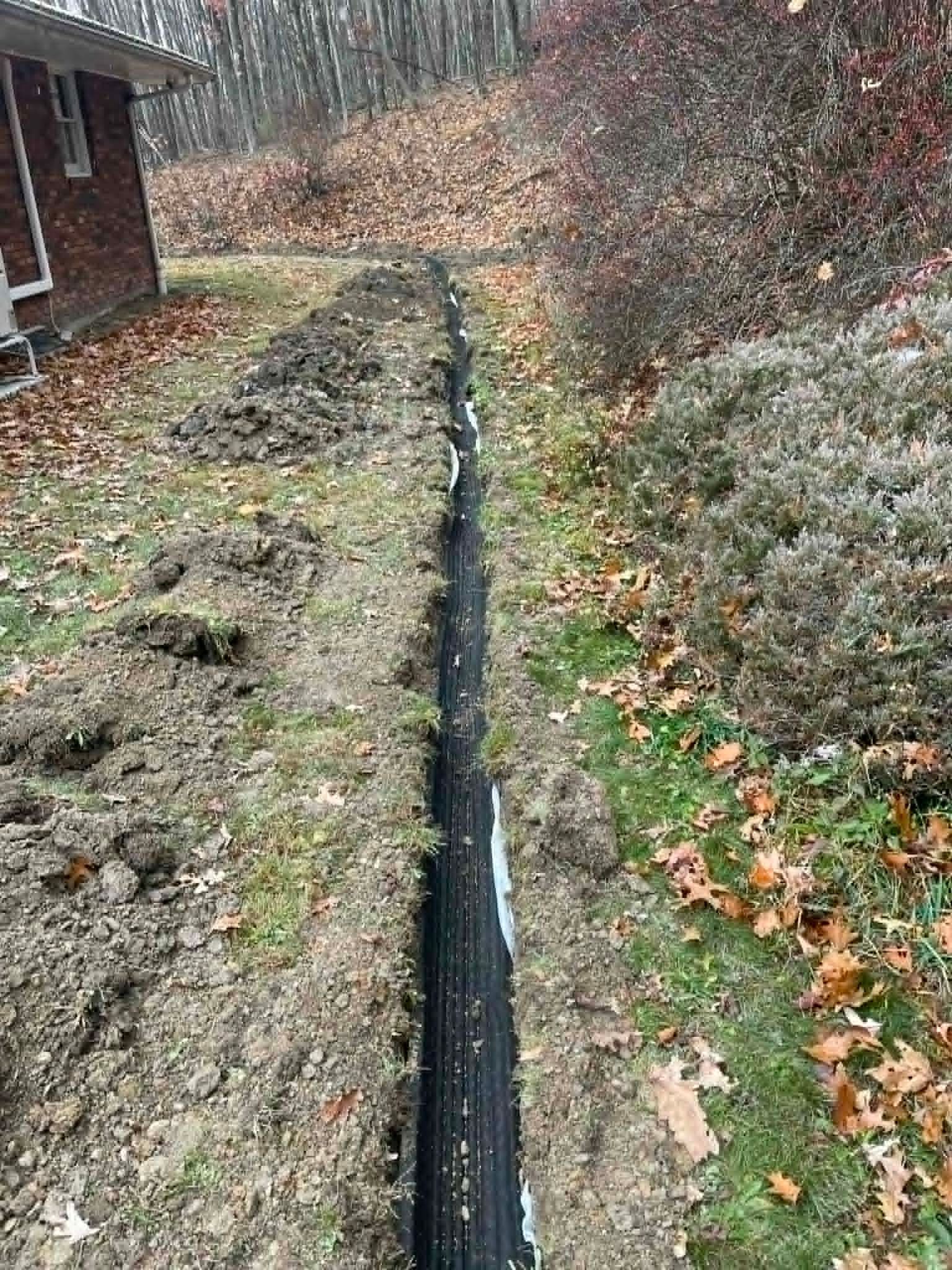 Narrow drainage trench with black liner beside a house and wooded slope, lined with fallen leaves and dirt sides