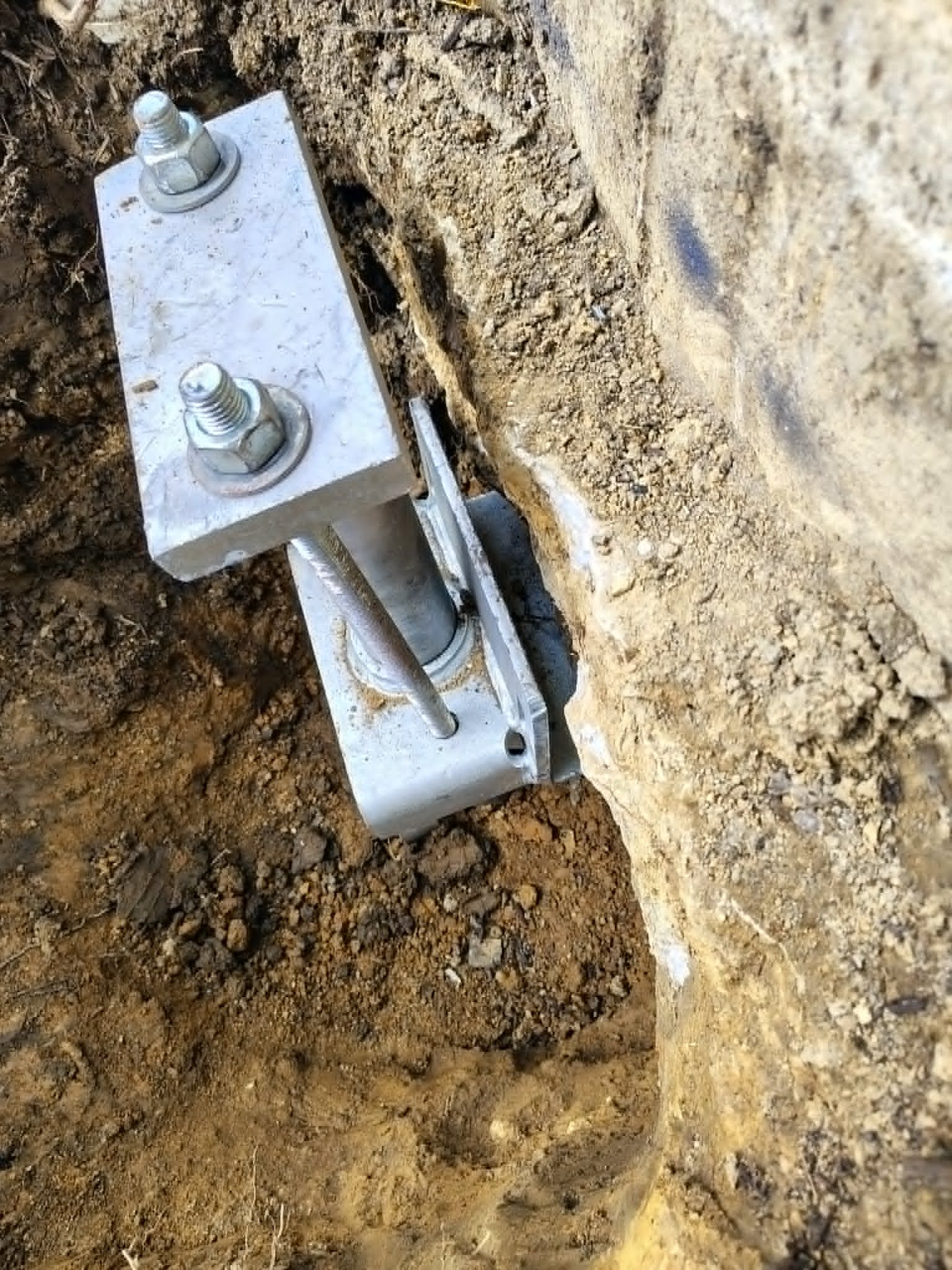 Close-up of a metal hinge or bracket bolted into dirt beside a concrete wall.