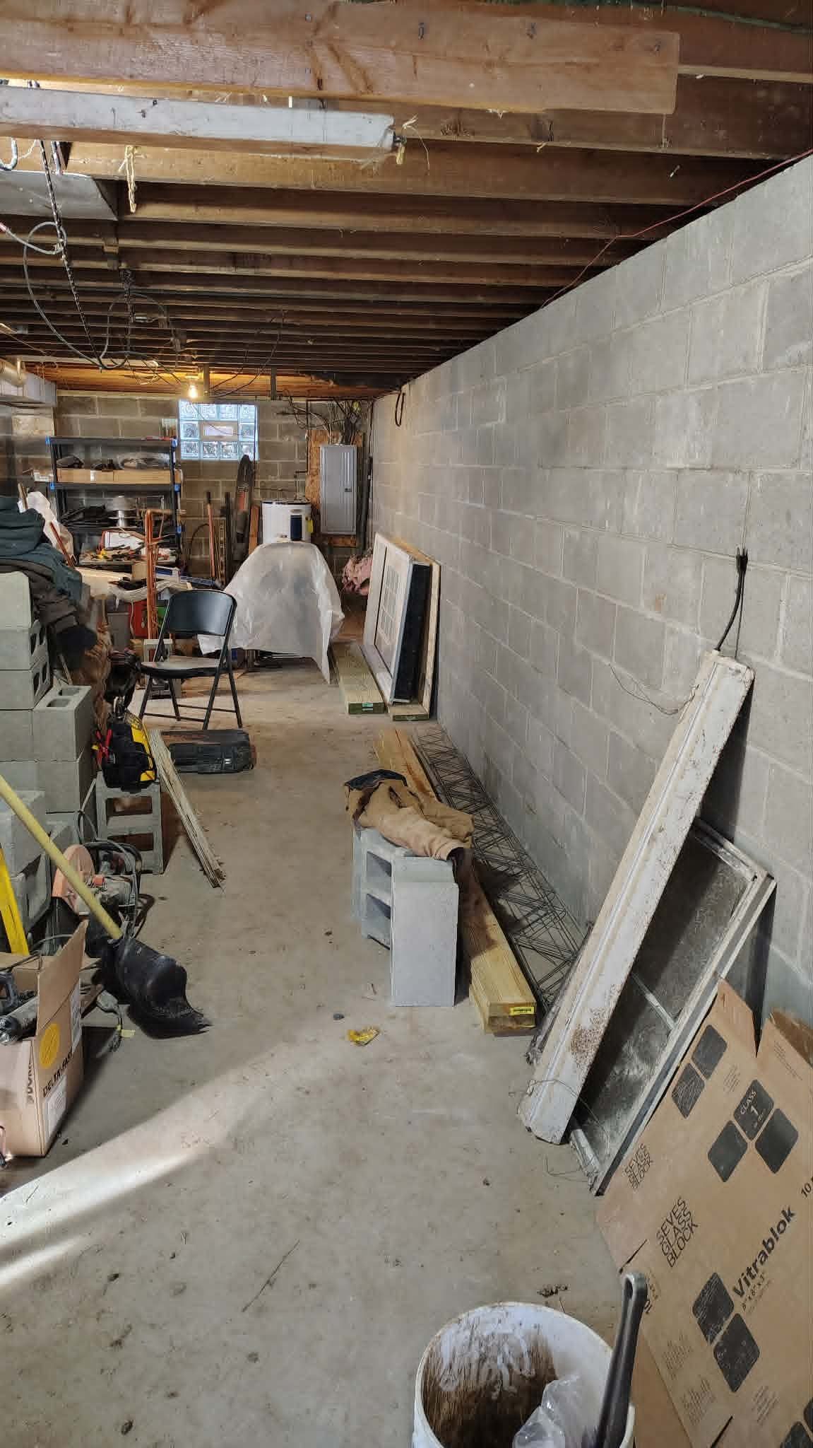 Cluttered basement workshop with tools, shelves, and a long cinder block wall under exposed ceiling joists