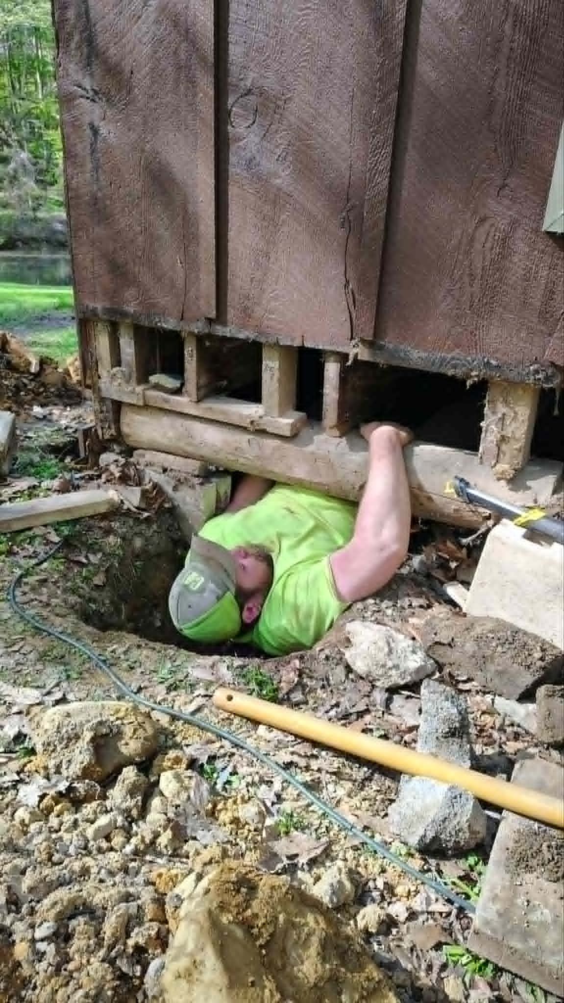 Person in a green shirt crawling under a raised wooden structure amid rubble and dirt.