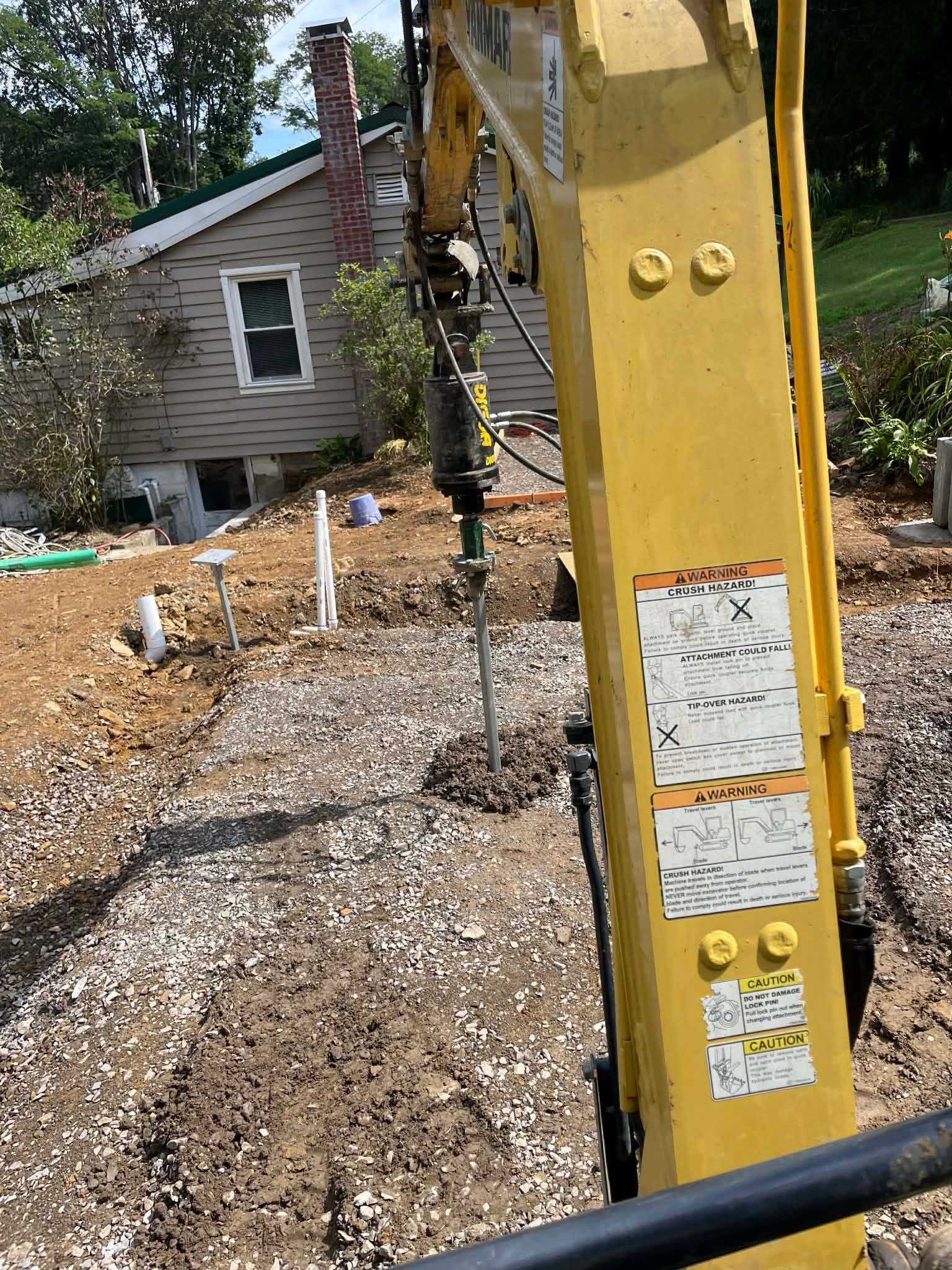 Yellow construction machinery arm in foreground at a gravel worksite beside a small house and trees.