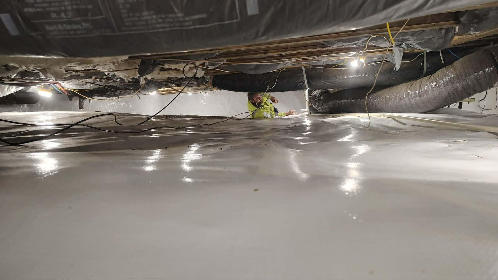 Inside a crawl space, plastic sheeting reflects light beneath exposed pipes and insulation.