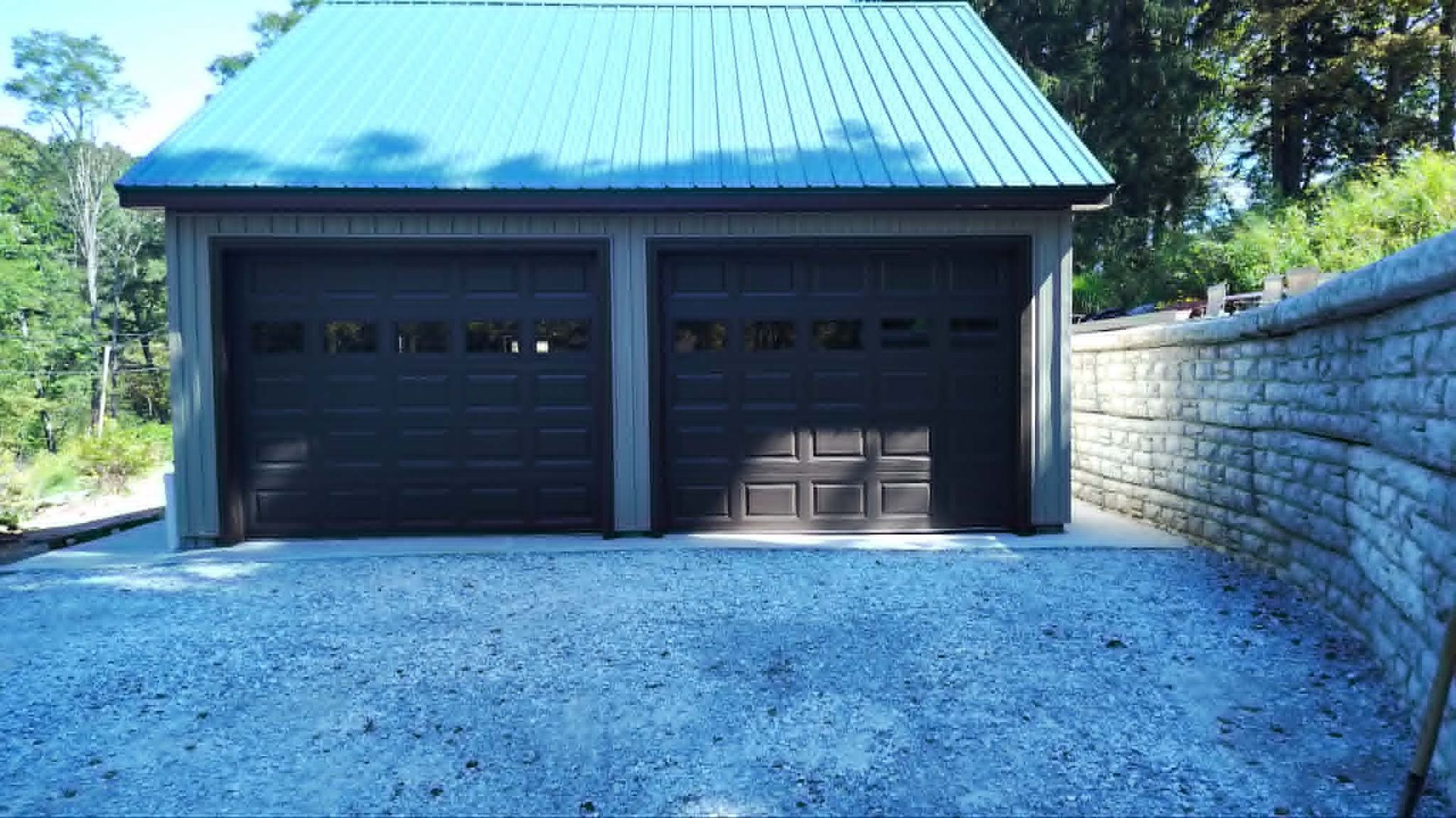 Double garage with dark doors, blue-green metal roof, and gravel driveway beside a stone retaining wall.