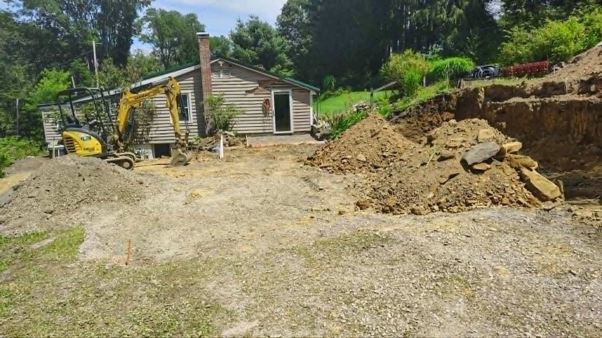 Yellow excavator at a dirt construction site beside a small building and hillside vegetation