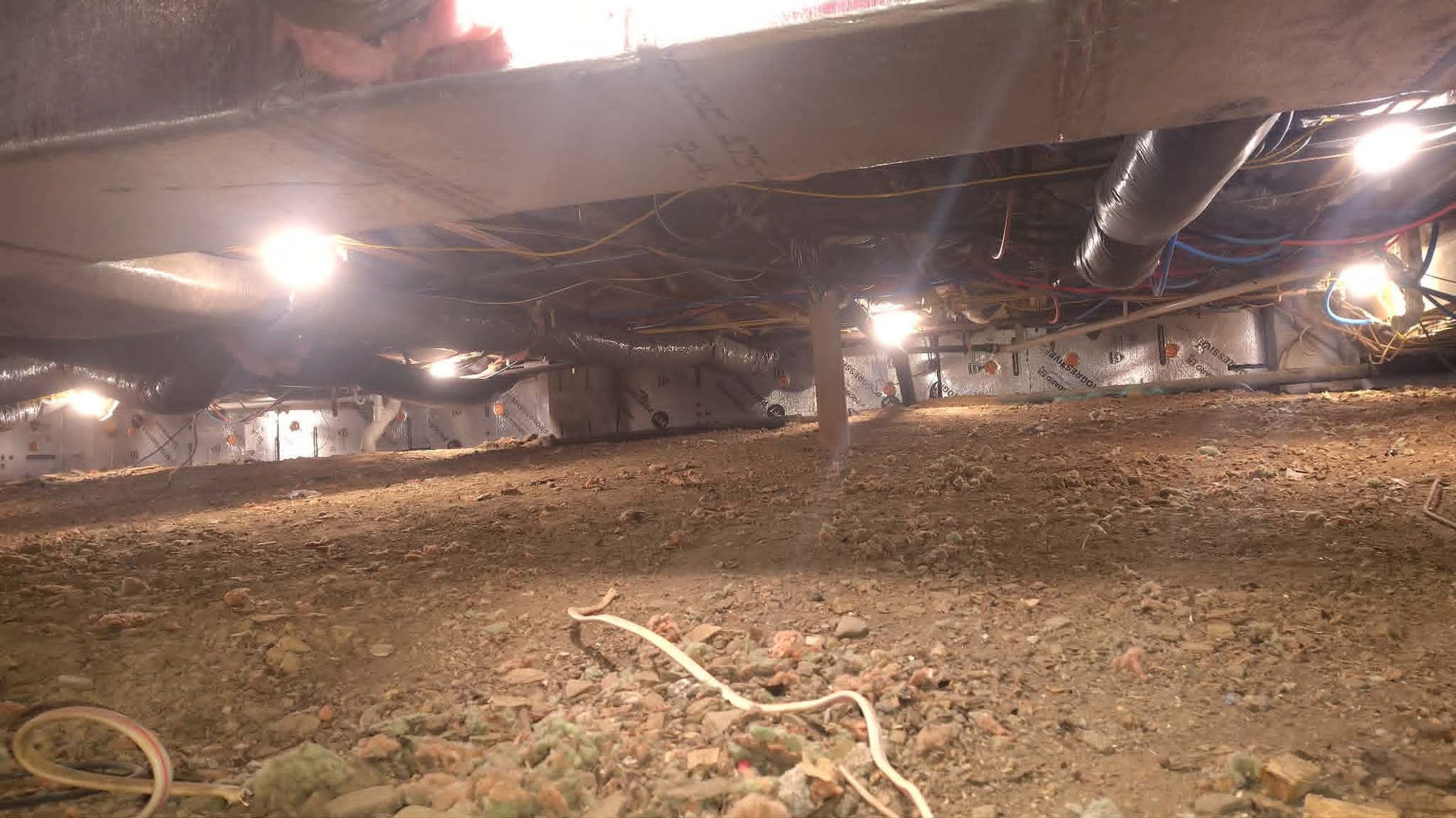 Crawlspace under a building with dirt floor, exposed pipes, beams, and hanging lights.
