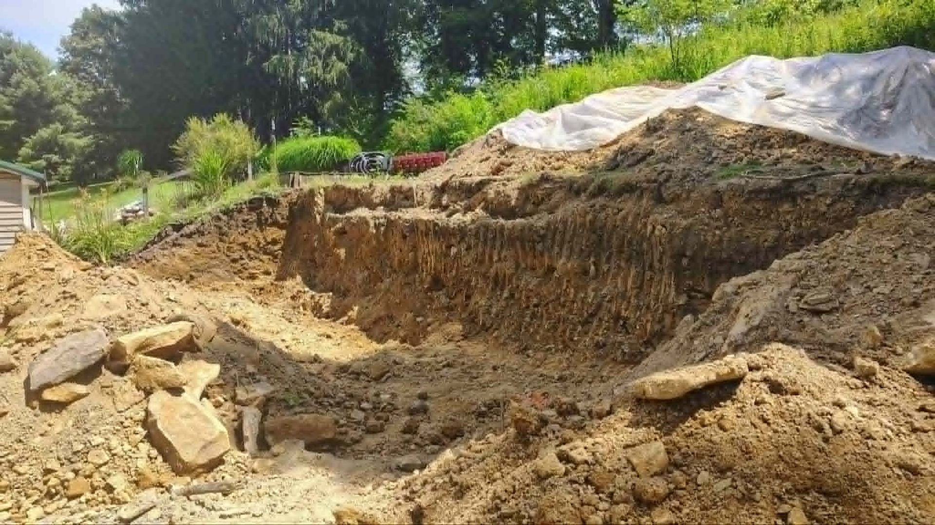Excavated dirt slope with tarp-covered soil piles in a wooded outdoor area