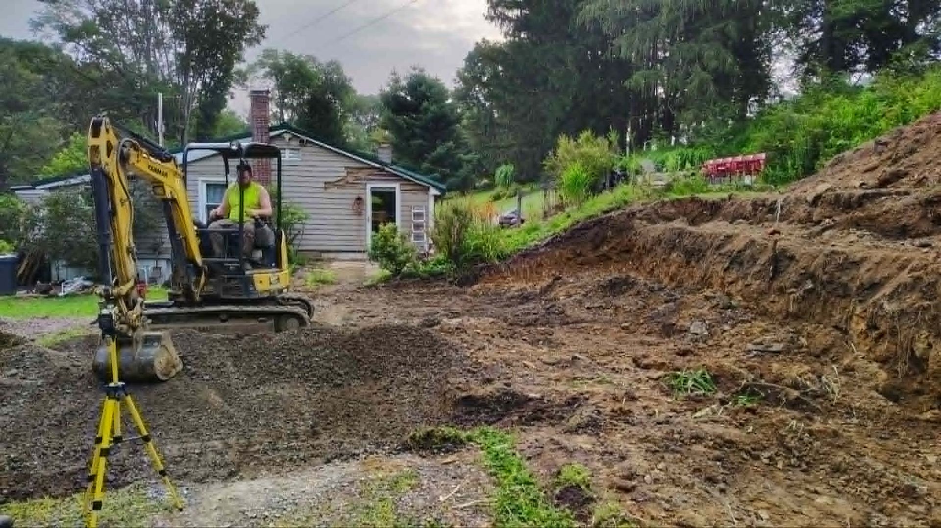 Excavator clearing a muddy hillside beside a small house after heavy rain