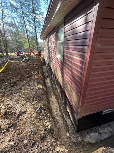 Red house siding over exposed concrete foundation beside a dirt trench and wooded yard